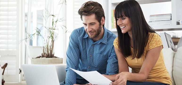 Man and woman sitting on couch looking at a laptop computer; woman is holding a sheet of paper