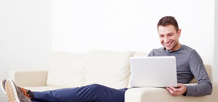 Man relaxing on a white couch while looking at his laptop computer.