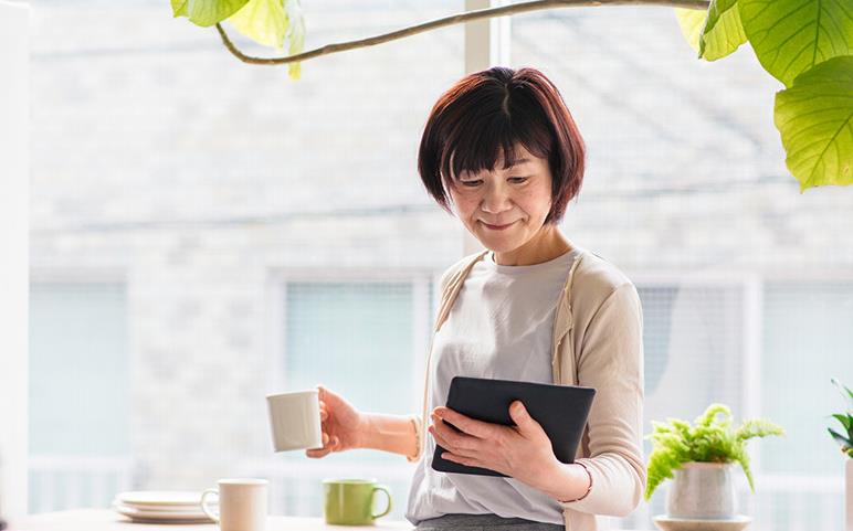A woman standing by a window, holding a cup of coffee, looking at a tablet device