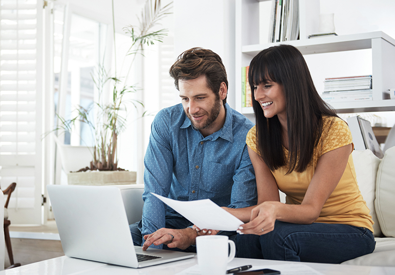 Man and woman sitting on couch looking at a laptop computer; woman is holding a sheet of paper