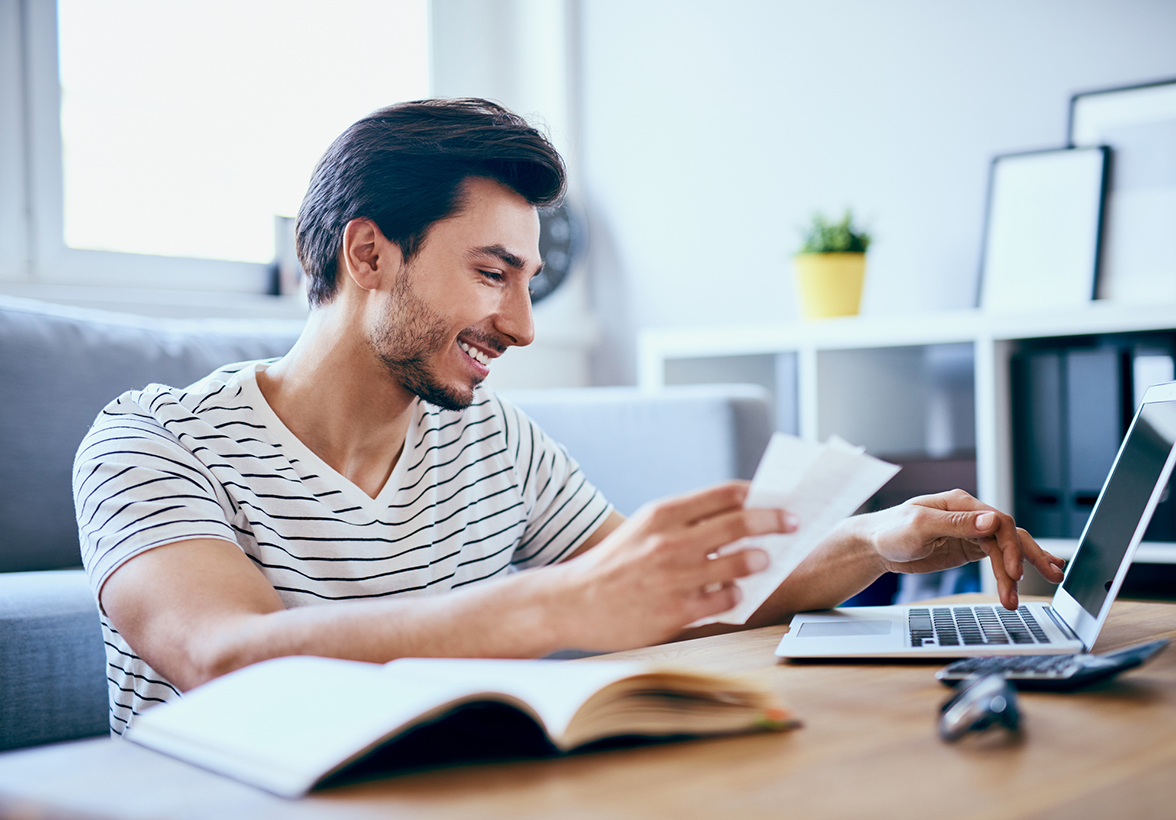Smiling young man sitting at a table paying bills on a laptop computer