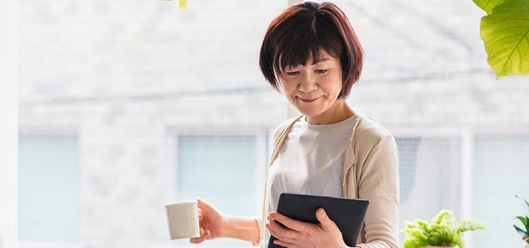 A woman standing by a window, holding a cup of coffee, looking at a tablet device
